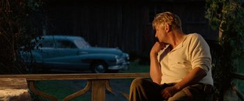 Movie still from “Under the Sun” (1998), directed by Colin Nutley – A man sitting on top of a bench next to an old car; Medium shot, Low angle
