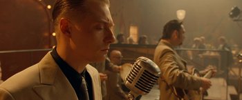 Movie still from “Under the Sun” (1998), directed by Colin Nutley – A man in a suit and tie is singing into a microphone; Close Up shot, Over the shoulder angle