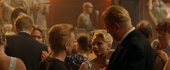 Movie still from “Under the Sun” (1998), directed by Colin Nutley – A group of people standing in a room talking to each other; Medium shot, Over the shoulder angle