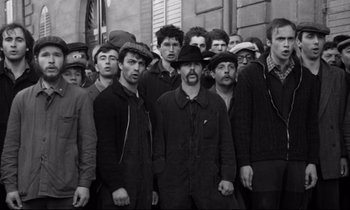 Movie still from “A Room in Town” (1982), directed by Jacques Demy – Black and white photograph of a group of men standing in front of a building; Medium shot, High angle