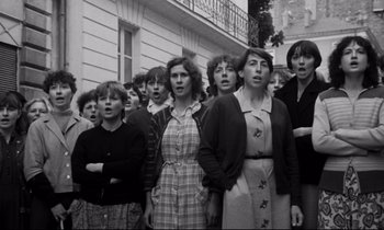 Movie still from “A Room in Town” (1982), directed by Jacques Demy – A black and white photo of a group of people standing in front of a building; Medium shot, High angle