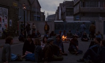 Movie still from “A Room in Town” (1982), directed by Jacques Demy – A group of people sitting on the ground next to a fire pit; Wide shot, High angle