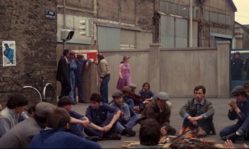 Movie still from “A Room in Town” (1982), directed by Jacques Demy – A group of people sitting on the ground near a fire pit; Wide shot, High angle