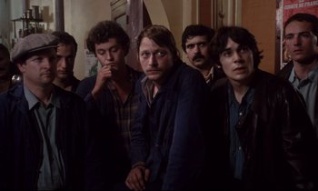 Movie still from “A Room in Town” (1982), directed by Jacques Demy – A group of men standing next to each other in a room; Medium shot, High angle