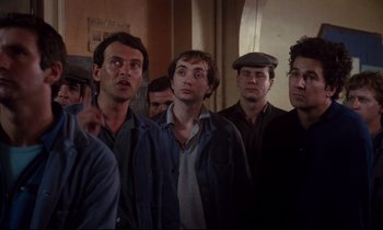 Movie still from “A Room in Town” (1982), directed by Jacques Demy – A group of men standing next to each other in a room; Medium shot, High angle