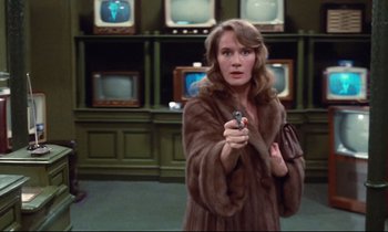 Movie still from “A Room in Town” (1982), directed by Jacques Demy – A woman holding a gun in a room full of tvs; Medium shot, Over the shoulder angle