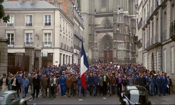 Movie still from “A Room in Town” (1982), directed by Jacques Demy – A large group of people standing in front of a building; Extreme Wide shot, High angle