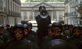 Movie still from “A Room in Town” (1982), directed by Jacques Demy – A group of men in helmets are standing in front of a building; Wide shot, High angle