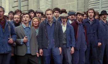 Movie still from “A Room in Town” (1982), directed by Jacques Demy – A group of men standing next to each other in front of a brick wall; Medium shot, High angle