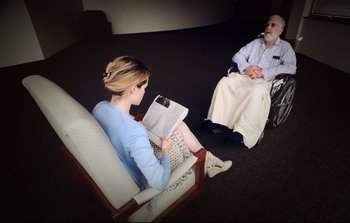 Movie still from “Unsane” (2018), directed by Steven Soderbergh – A woman sitting in a chair reading a book to an older man in a wheel chair; Medium shot, Over the shoulder angle