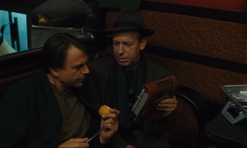 Movie still from “Until the End of the World” (1991), directed by Wim Wenders – Two men sitting next to each other holding an orange; Medium shot, High angle