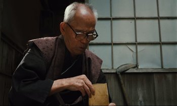 Movie still from “Until the End of the World” (1991), directed by Wim Wenders – An older man wearing glasses and holding a piece of wood; Medium shot, Low angle