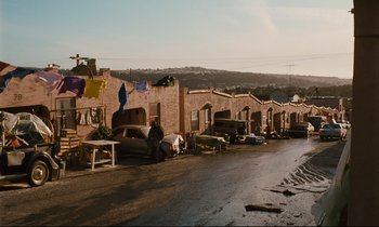 Movie still from “Until the End of the World” (1991), directed by Wim Wenders – A man sitting on the side of the road next to a row of houses; Extreme Wide shot, High angle