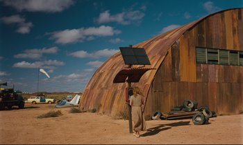 Movie still from “Until the End of the World” (1991), directed by Wim Wenders – A woman standing in front of an old barn holding a solar panel; Extreme Wide shot, Low angle