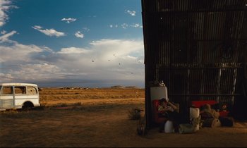 Movie still from “Until the End of the World” (1991), directed by Wim Wenders – A woman sitting on a chair next to an open gate; Extreme Wide shot, Low angle