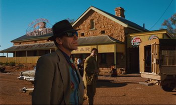 Movie still from “Until the End of the World” (1991), directed by Wim Wenders – A man in a suit and hat standing in front of a building; Wide shot, Over the shoulder angle