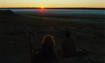 Movie still from “Until the End of the World” (1991), directed by Wim Wenders – Two people sitting in the grass watching the sun go down; Wide shot, High angle