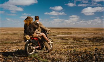 Movie still from “Until the End of the World” (1991), directed by Wim Wenders – A man and a woman riding a bike through a field; Wide shot, High angle