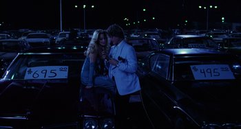 Movie still from “Used Cars” (1980), directed by Robert Zemeckis – A man and a woman standing next to each other in a parking lot at night; Wide shot, Over the shoulder angle