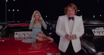 Movie still from “Used Cars” (1980), directed by Robert Zemeckis – A woman sitting on the hood of a red car next to a man; Medium shot, Low angle