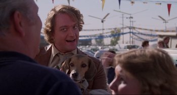 Movie still from “Used Cars” (1980), directed by Robert Zemeckis – A man holding a brown and white dog in his arms; Close Up shot, Over the shoulder angle