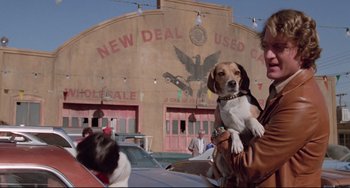 Movie still from “Used Cars” (1980), directed by Robert Zemeckis – A woman holding a brown and white dog in front of a building; Medium shot, Low angle