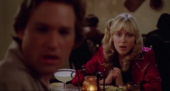 Movie still from “Used Cars” (1980), directed by Robert Zemeckis – A woman sitting at a table with a man in the background; Close Up shot, Over the shoulder angle