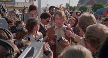 Movie still from “Used Cars” (1980), directed by Robert Zemeckis – A crowd of people gathered around a man in a brown suit; Medium shot, Over the shoulder angle