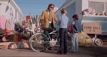 Movie still from “Used Cars” (1980), directed by Robert Zemeckis – A group of people standing next to a bicycle; Wide shot, Over the shoulder angle