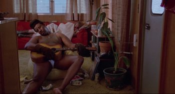 Movie still from “Used Cars” (1980), directed by Robert Zemeckis – A man sitting on a couch playing a guitar; Wide shot, High angle