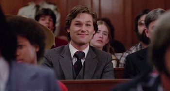Movie still from “Used Cars” (1980), directed by Robert Zemeckis – A man in a suit and tie sitting in front of a group of people; Close Up shot, Over the shoulder angle