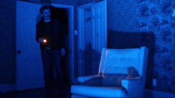 Movie still from “V/H/S” (2012), directed by Joe Swanberg – A man standing in a dark room holding a flashlight; Wide shot, High angle