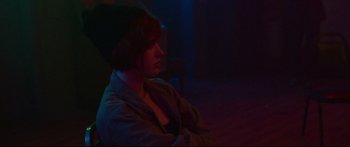 Movie still from “VFW” (2019), directed by Joe Begos – A woman sitting in a dark room with her arms folded on her chest; Medium shot, Over the shoulder angle