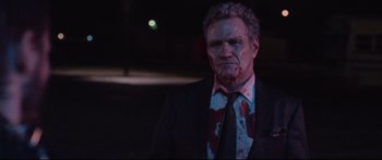 Movie still from “VFW” (2019), directed by Joe Begos – A man in a suit and tie with blood all over his face; Medium shot, Over the shoulder angle