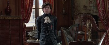 Movie still from “Valmont” (1989), directed by Milos Forman – A young man in a suit stands next to a chair in a room; Medium shot, Over the shoulder angle