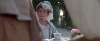 Movie still from “Valmont” (1989), directed by Milos Forman – A woman wearing a hat and a dress sitting at an outdoor table; Medium shot, Over the shoulder angle