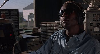 Movie still from “Vanishing Point” (1971), directed by Richard C. Sarafian – A man wearing sunglasses sitting in front of a stack of books; Close Up shot, Low angle