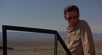 Movie still from “Vanishing Point” (1971), directed by Richard C. Sarafian – A man in a sheriff's uniform standing next to a vehicle; Medium shot, Low angle