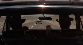 Movie still from “Vanishing Point” (1971), directed by Richard C. Sarafian – A car driving down a road with a person in the back seat of the car; Wide shot, Over the shoulder angle