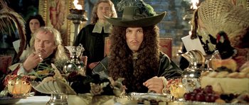 Movie still from “Vatel” (2000), directed by Roland Joffé – A man sitting at a dinner table wearing a hat; Medium shot, Over the shoulder angle