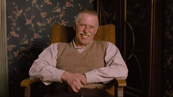 Movie still from “Vera Drake” (2004), directed by Mike Leigh – An older man sitting in a chair smiling for the camera; Medium shot, Low angle
