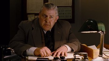 Movie still from “Vera Drake” (2004), directed by Mike Leigh – An older man sitting at a desk in front of papers; Medium shot, Low angle