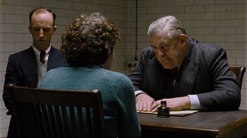 Movie still from “Vera Drake” (2004), directed by Mike Leigh – An older man sitting at a table talking to two other people; Medium shot, Over the shoulder angle
