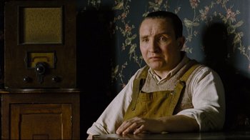 Movie still from “Vera Drake” (2004), directed by Mike Leigh – A man sitting at a table in front of a floral wallpaper; Medium shot, Low angle