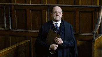 Movie still from “Vera Drake” (2004), directed by Mike Leigh – An older man in a suit and tie holding a folder; Medium shot, Low angle