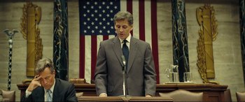 Movie still from “Vice” (2018), directed by Adam McKay – A man standing at a podium in front of an american flag; Medium shot, Low angle