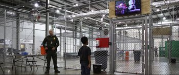 Movie still from “Vice” (2018), directed by Adam McKay – A young boy standing in front of a fence watching a television screen; Wide shot, Low angle