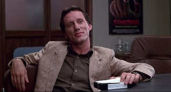 Movie still from “Videodrome” (1983), directed by David Cronenberg – A man sitting at a table with a book in his hand; Close Up shot, Low angle