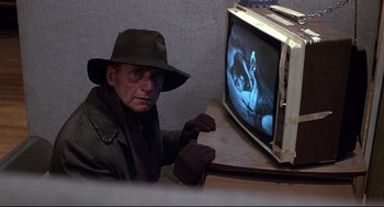 Movie still from “Videodrome” (1983), directed by David Cronenberg – A man wearing a hat and a scarf sitting in front of a tv; Close Up shot, High angle