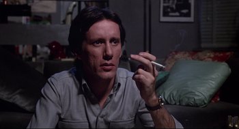 Movie still from “Videodrome” (1983), directed by David Cronenberg – A man sitting on a couch holding a cigarette in his hand; Close Up shot, Low angle